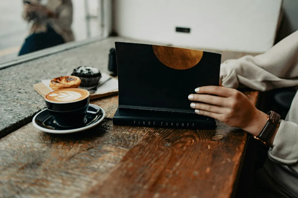 A hand resting on a dark notebook beside a cappuccino on a worn wooden table