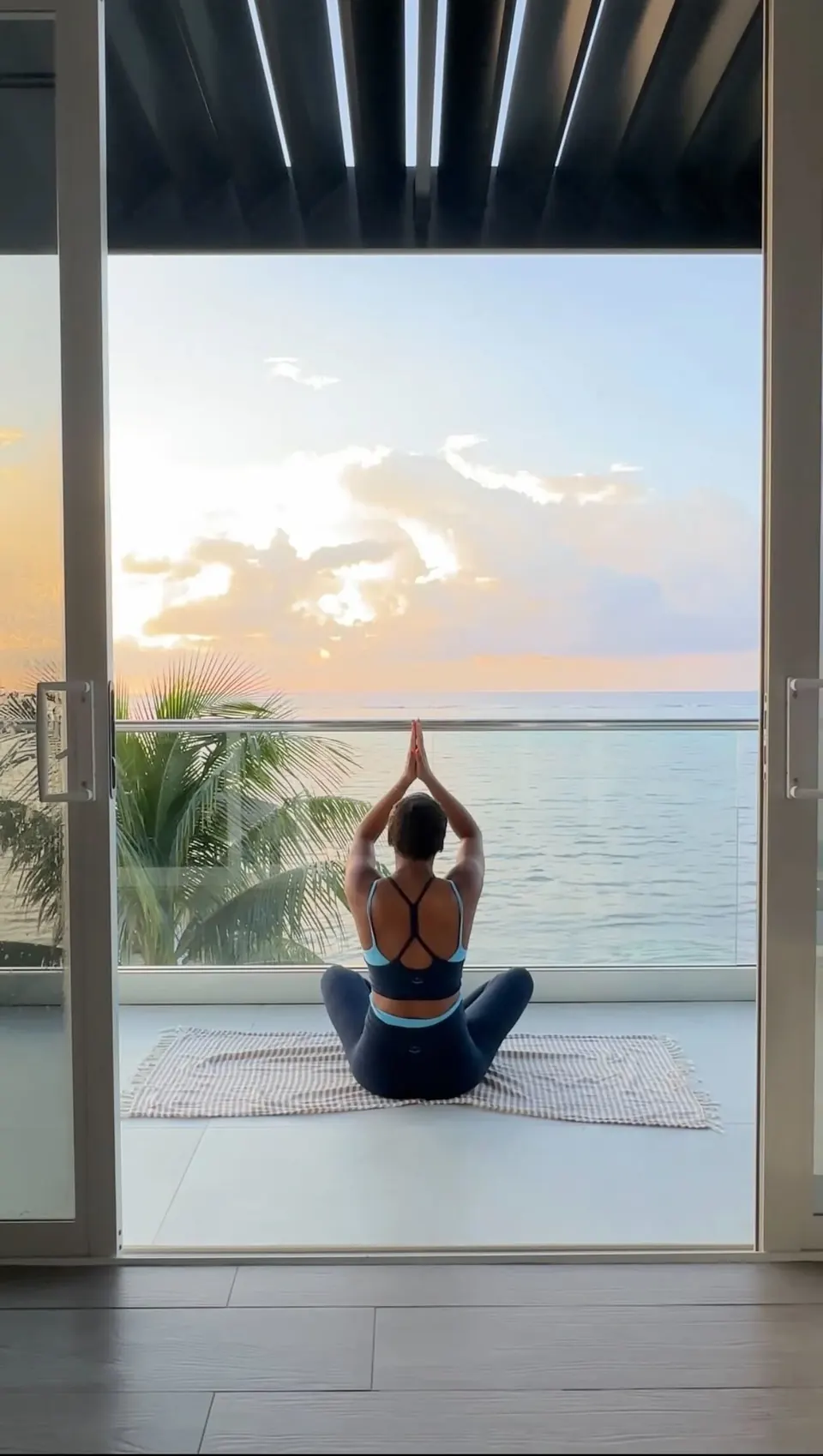 A woman seated cross-legged with hands overhead, facing an open balcony and the ocean at sunrise